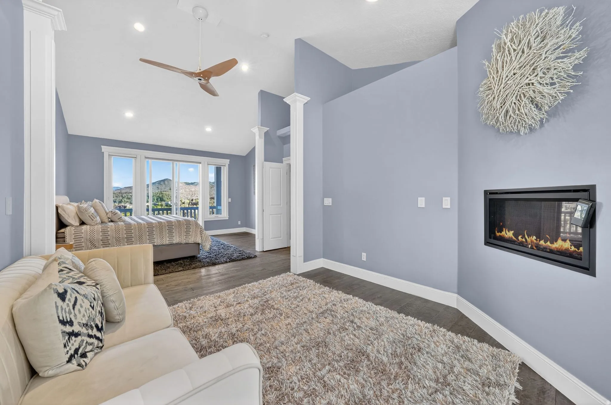 Bedroom with vaulted ceiling, a glass covered fireplace, decorative columns, dark wood-style flooring, and a ceiling fan