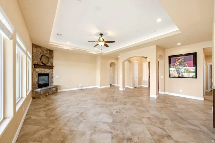 Unfurnished living room with arched walkways, a stone fireplace, ceiling fan, a raised ceiling, and recessed lighting