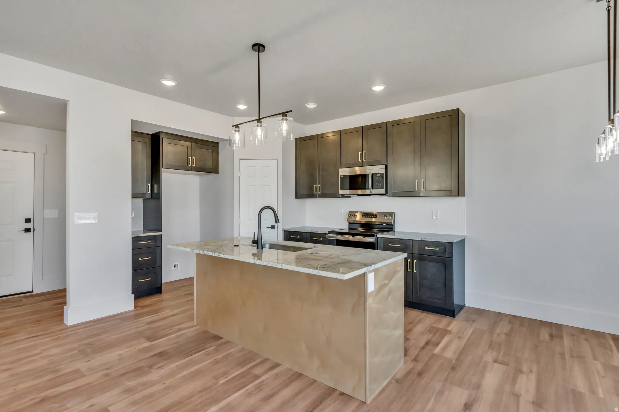 Kitchen featuring stainless steel appliances, light stone counters, pendant lighting, light wood-style flooring, and an island with sink
