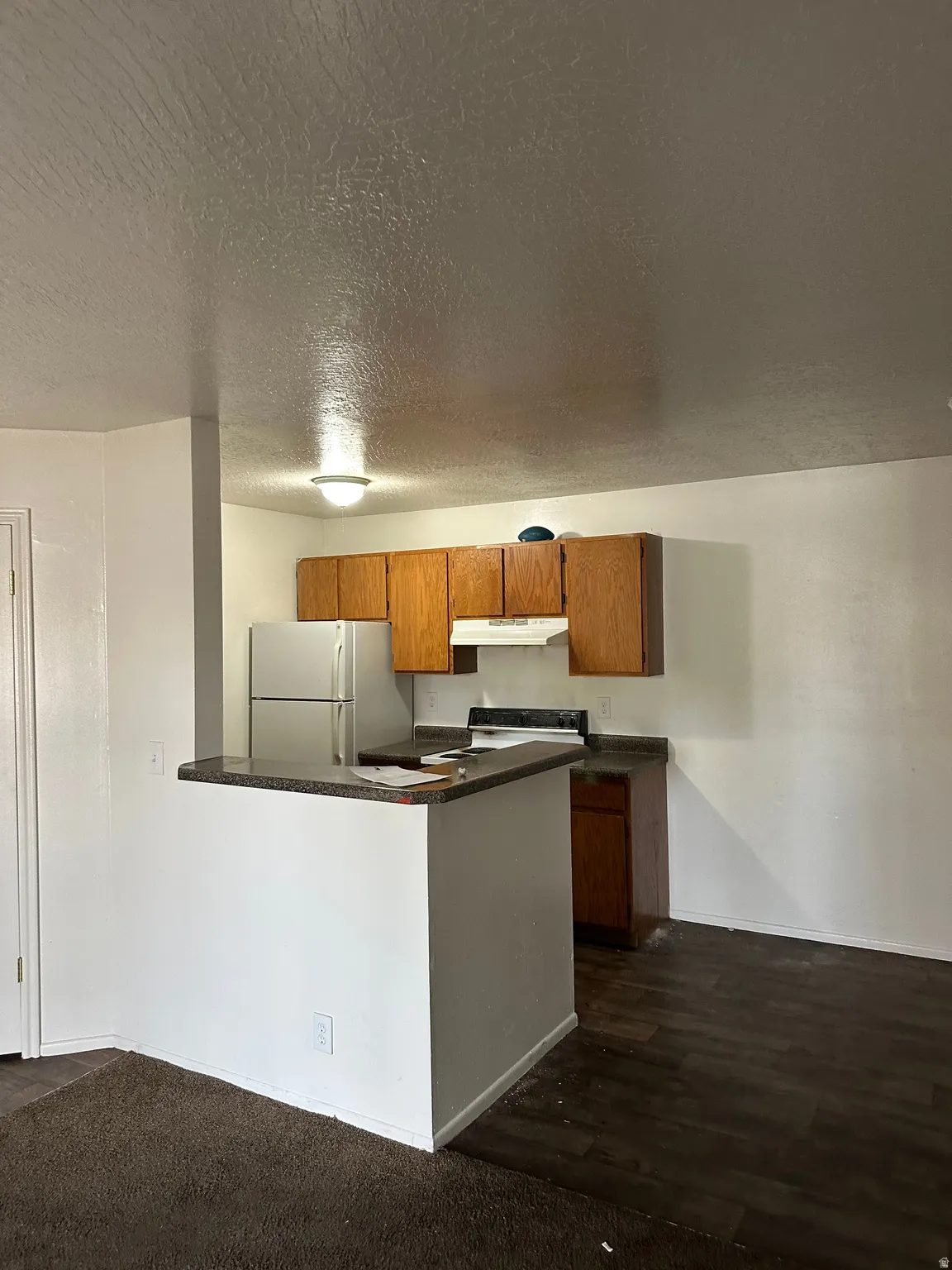 Kitchen featuring wood finish cabinetry, dark countertops, white appliances, a peninsula, and a textured ceiling