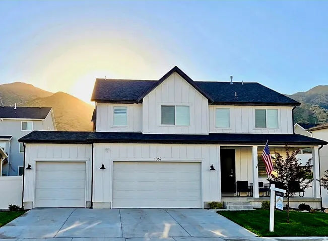 Modern farmhouse-style home featuring white vertical and horizontal siding, a dark shingle roof, two garage doors, a covered front porch, and a mountain backdrop