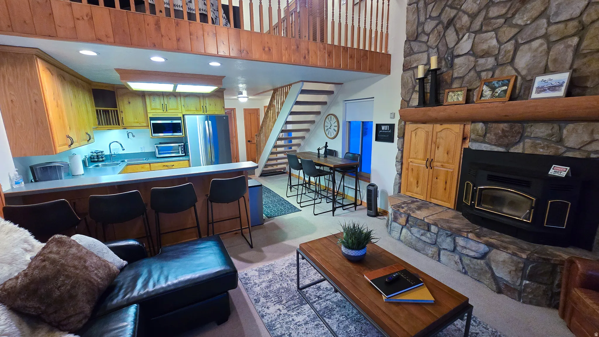 Open-concept living area featuring a stone-clad fireplace, wood-finish flooring, and a kitchen with wood cabinetry and stainless steel appliances