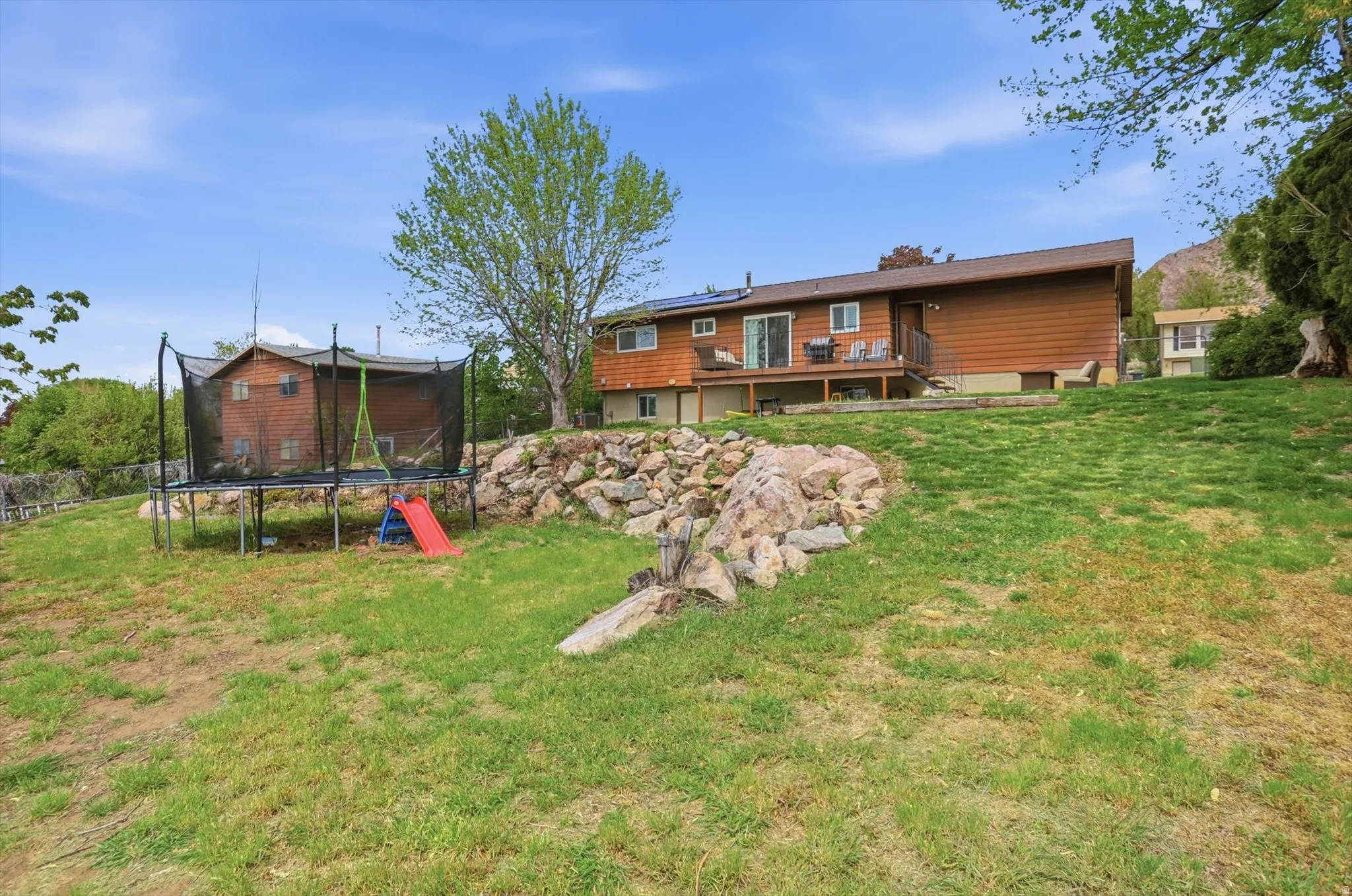 Back of property featuring a trampoline, a deck, a lawn, roof mounted solar panels, and a playground
