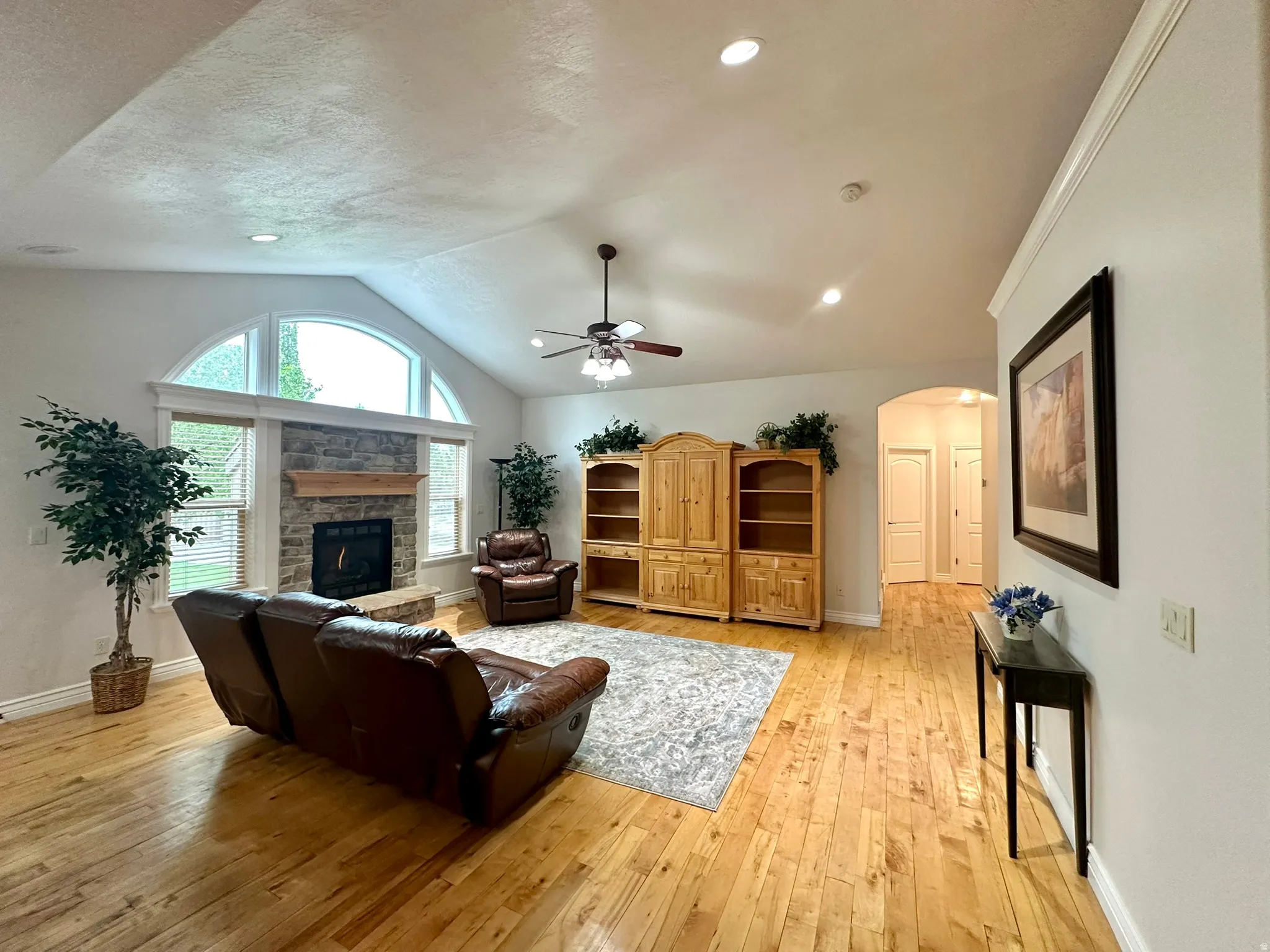 Vaulted ceiling with recessed lighting and ceiling fan, hard wood-finish flooring, stone-faced fireplace with wood mantel, arched window, and white trim throughout