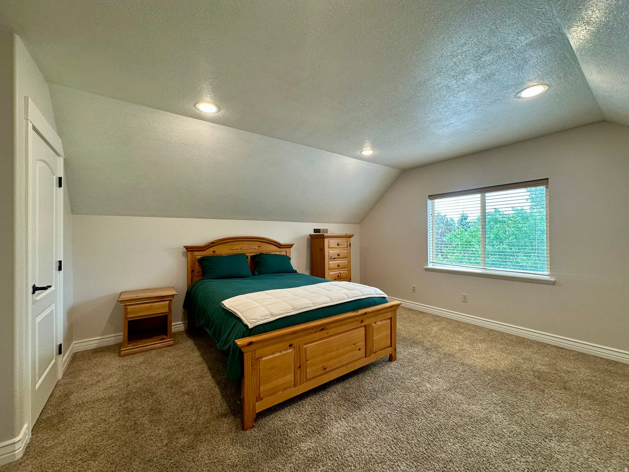 Spacious room featuring vaulted ceilings with recessed lighting, a large window with blinds, light-colored carpeting and dual walk-in closets.