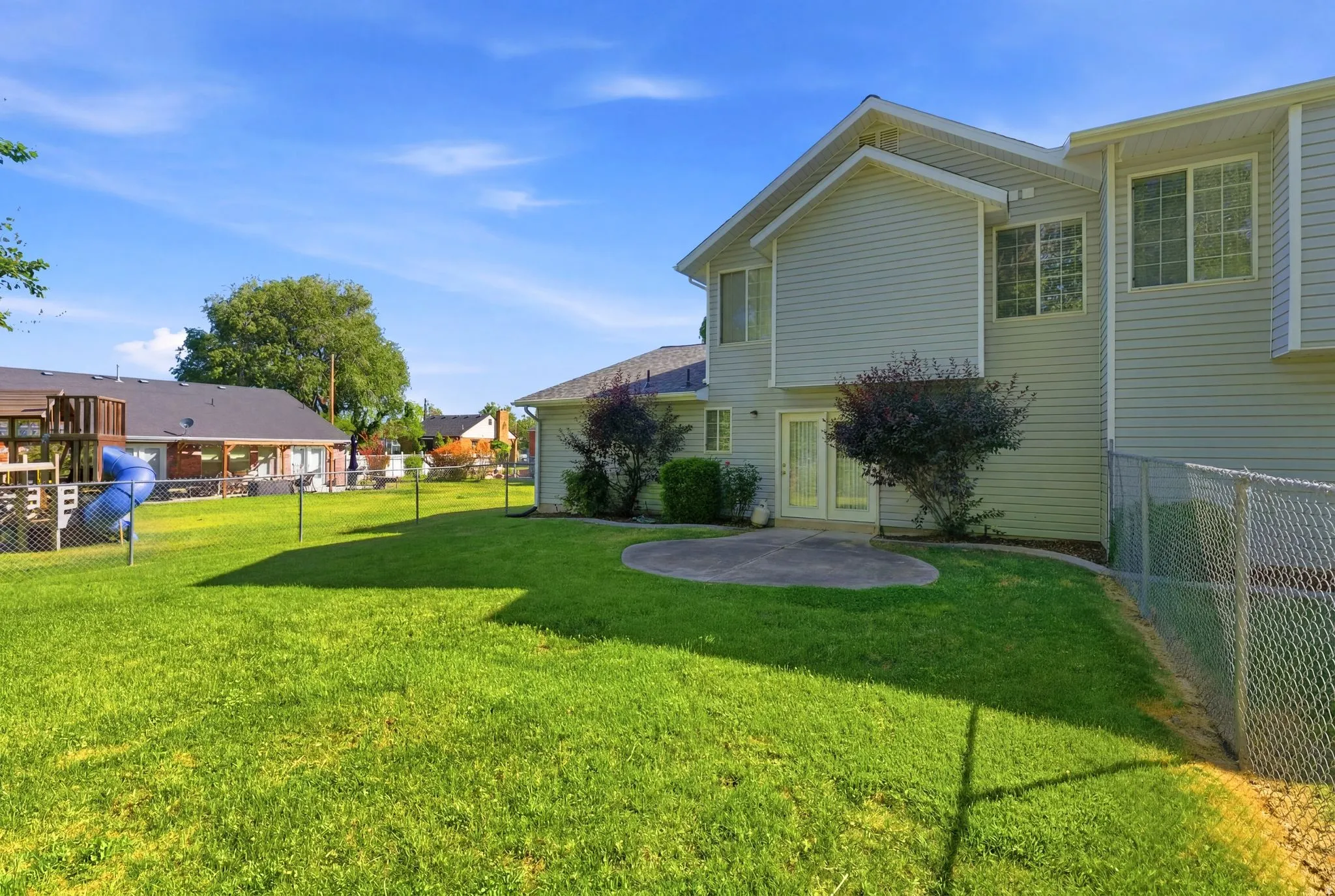 Expansive rear yard featuring green lawn, established landscaping, a concrete patio, and a chain-link fence
