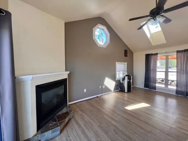 Vaulted ceiling with an installed ceiling fan, wood-finish flooring, corner fireplace with a tiled hearth, octagonal stained-glass window, and a skylight