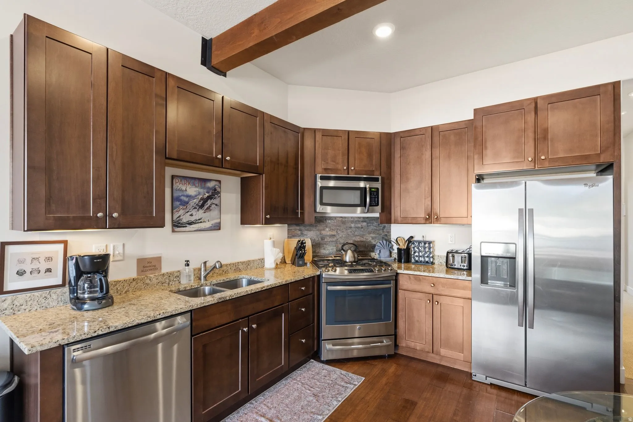Kitchen featuring stainless steel appliances, light stone countertops, backsplash, beam ceiling, and dark wood finish cabinetry
