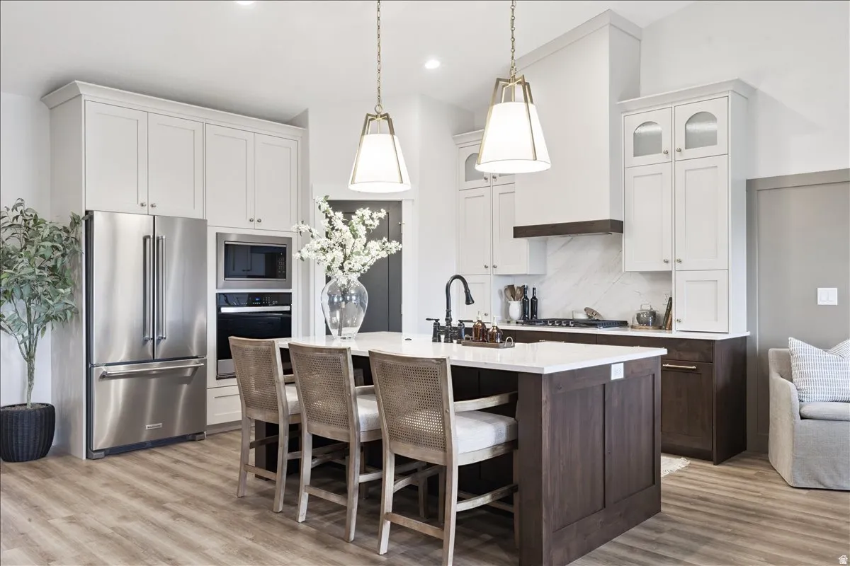Kitchen with a breakfast bar area, stainless steel appliances, a center island with sink, glass fronted cabinets, and light wood-style floors
