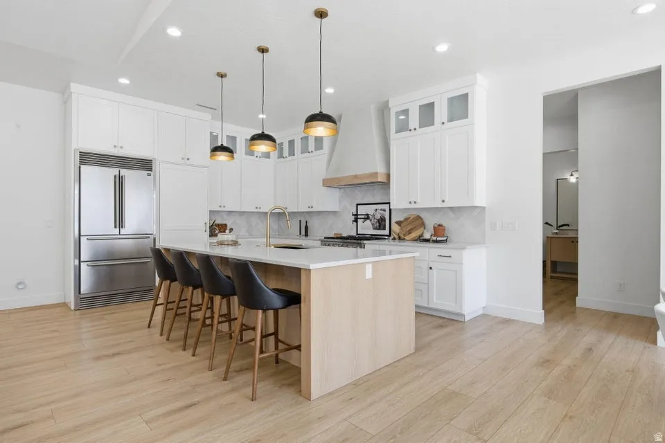 Two tone kitchen with hanging light fixtures, a kitchen island with sink, light wood-style flooring, built in fridge, and light stone counters
