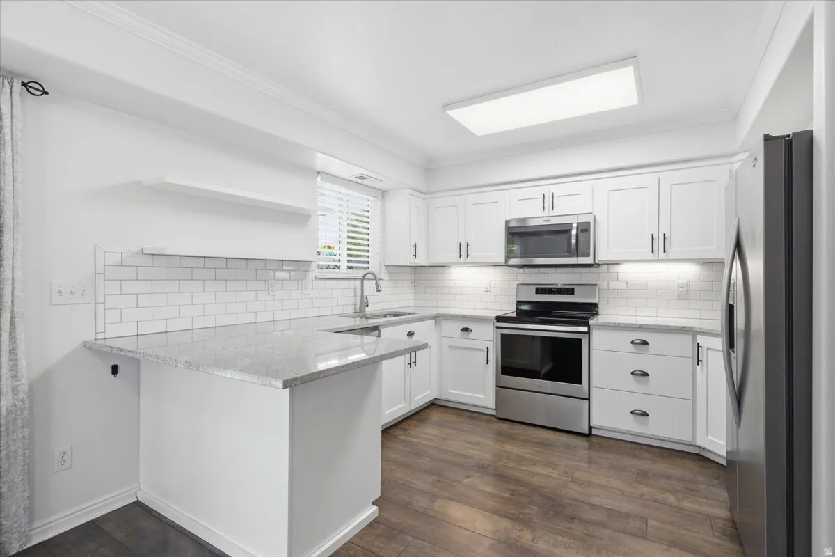 Kitchen with stainless steel appliances, ornamental molding, white cabinets, dark wood-type flooring, and backsplash