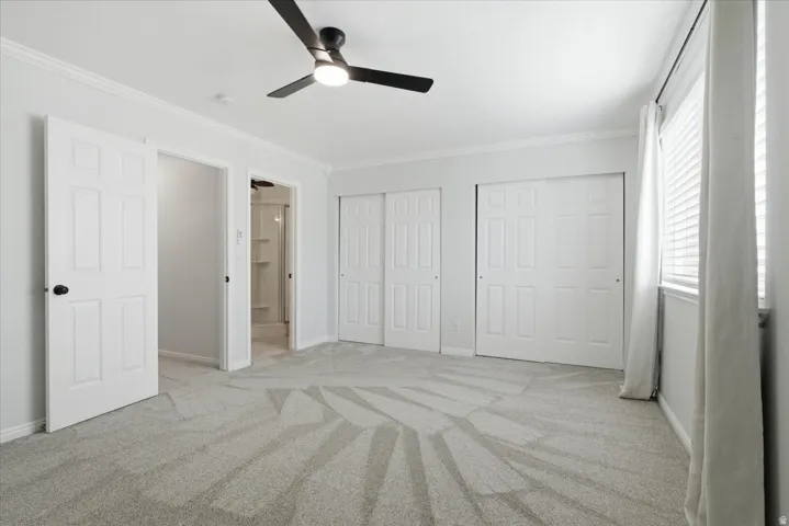Unfurnished bedroom with ensuite bathroom, multiple closets, light colored carpet, ornamental molding, and ceiling fan