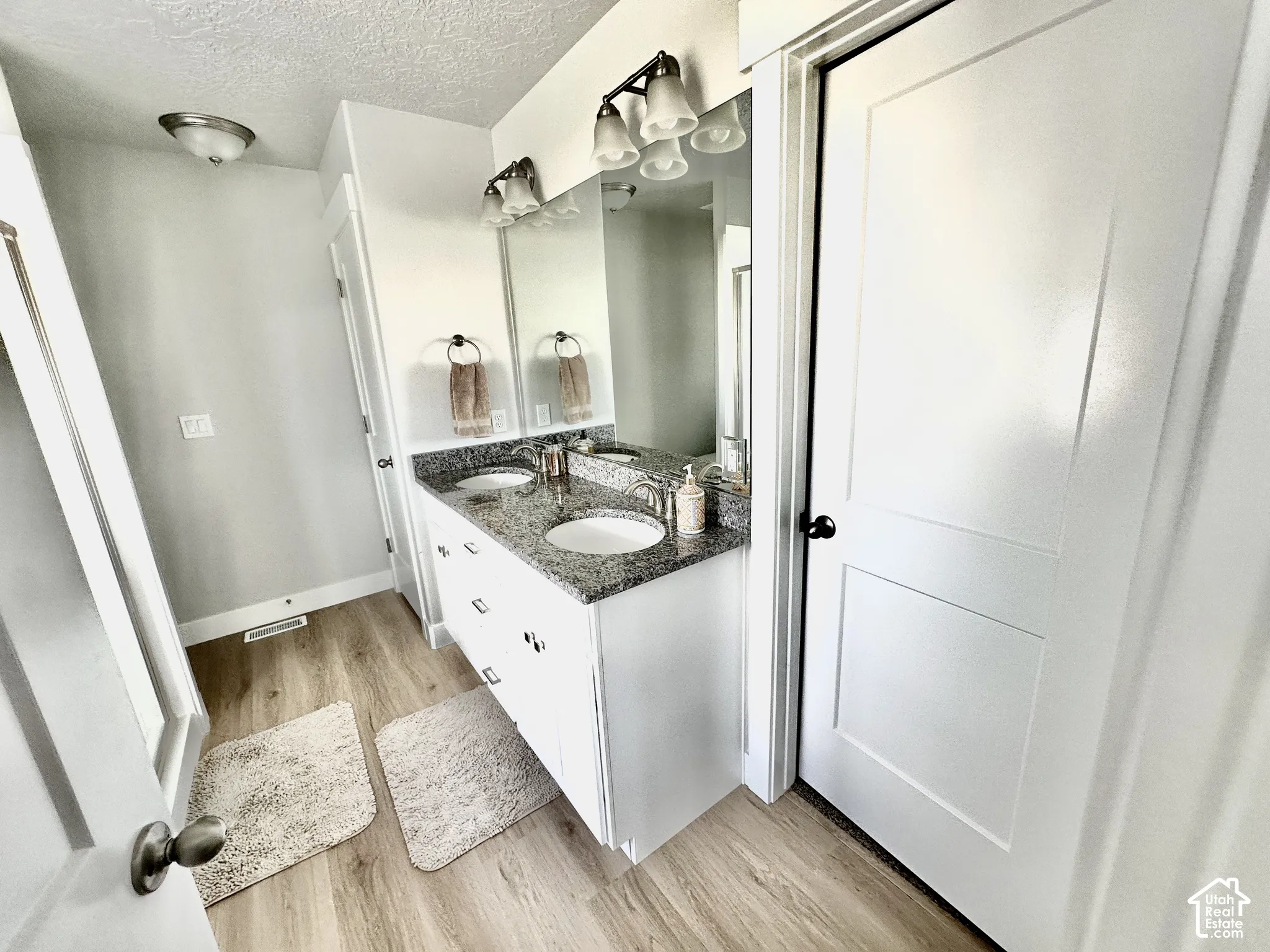 Double vanity with integrated sinks, granite-look countertop, white cabinetry, and wood-finish flooring