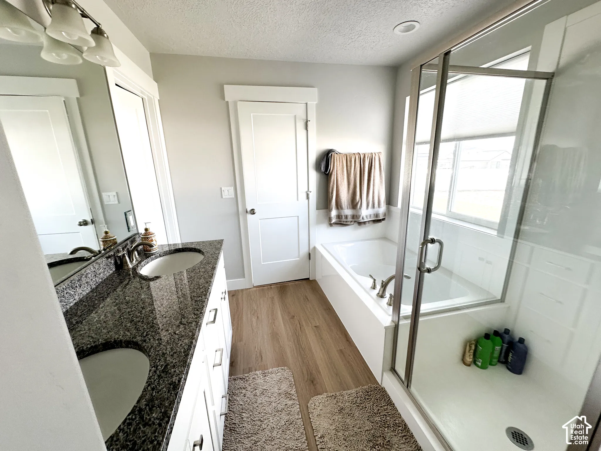 Dual vanity with dark granite countertops, built-in soaking tub, glass-enclosed shower, wood-finish flooring, and shaker-style cabinetry