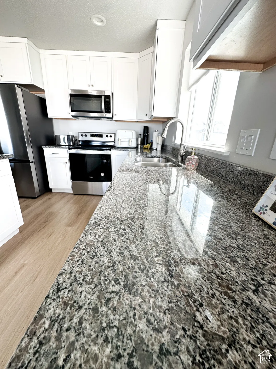 Granite countertops, white shaker-style cabinetry, stainless steel appliances, stainless steel sink with a gooseneck faucet, and wood-finish flooring