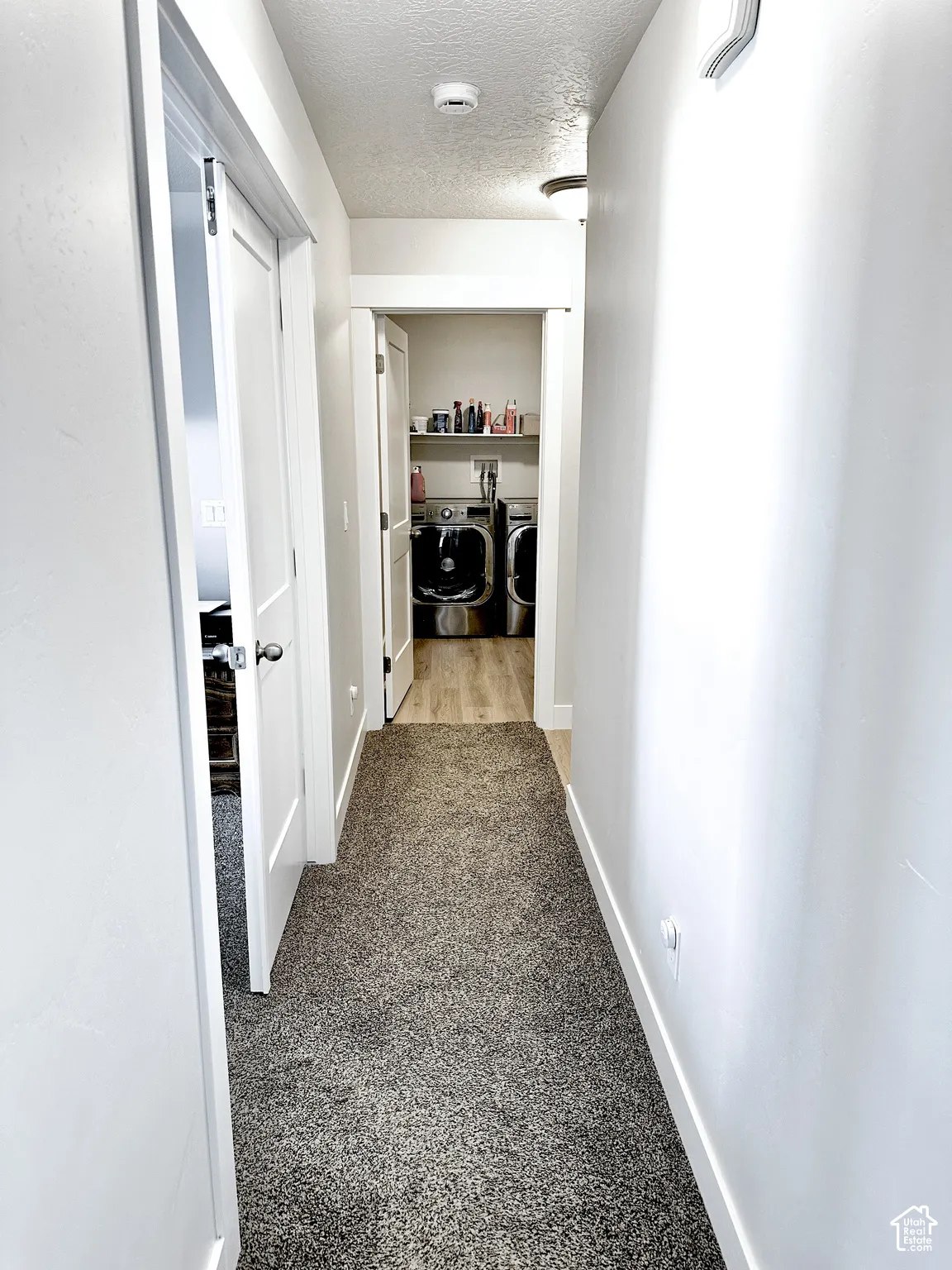 Hallway featuring carpet flooring and white walls, leading to a laundry area with wood-finish flooring and a built-in shelf
