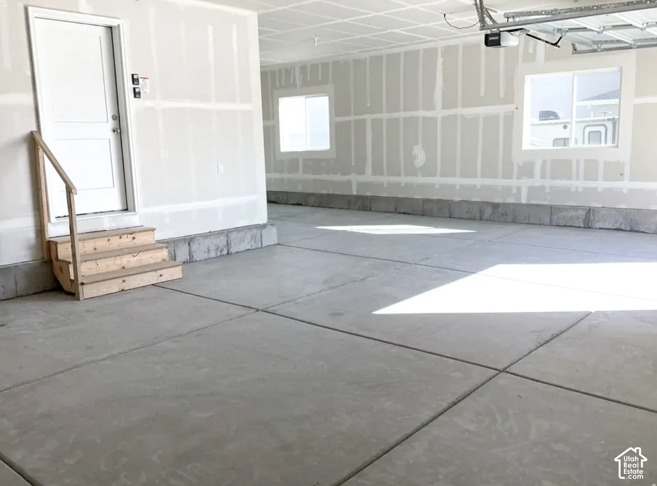 Concrete flooring with expansion joints, unfinished drywall, interior door with wood steps, two windows, and exposed garage door opener mechanism