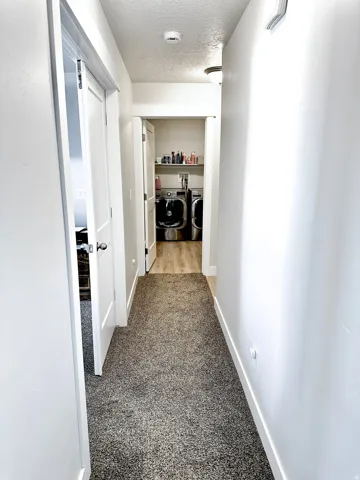 Hallway featuring carpet flooring and white walls, leading to a laundry area with wood-finish flooring and a built-in shelf
