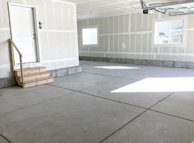 Concrete flooring with expansion joints, unfinished drywall, interior door with wood steps, two windows, and exposed garage door opener mechanism