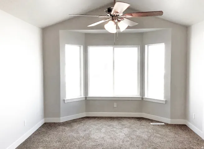 Vaulted ceiling, ceiling fan with light fixture, bay window, carpet flooring, and white trim