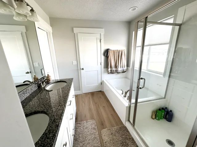 Dual vanity with dark granite countertops, built-in soaking tub, glass-enclosed shower, wood-finish flooring, and shaker-style cabinetry