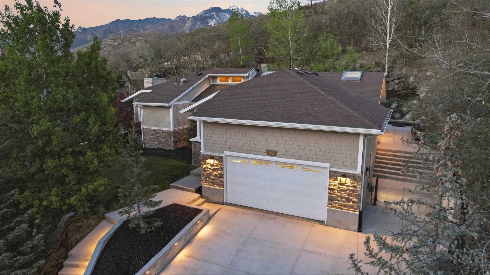 View of front of property featuring concrete driveway, stone siding, roof with shingles, a mountain view, and view of wooded area