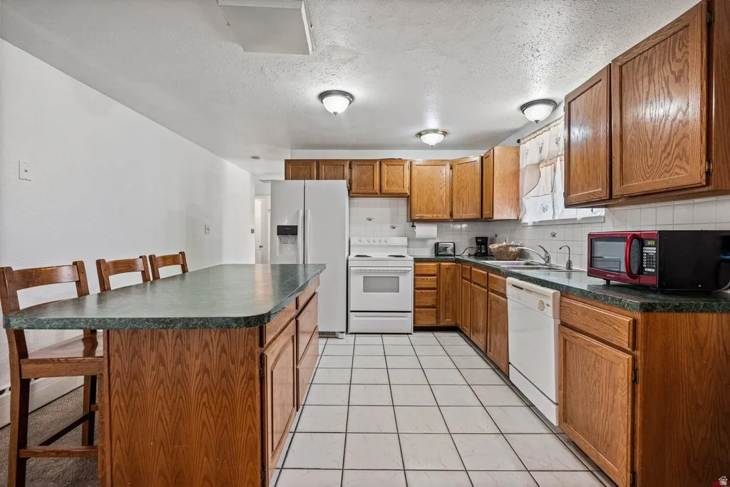 Kitchen with dark countertops, a kitchen bar, wood finish cabinetry, white appliances, and a textured ceiling