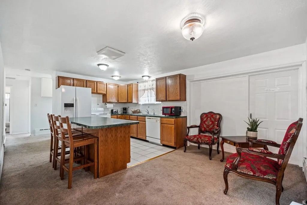 Kitchen featuring wood finish cabinetry, light colored carpet, dark countertops, white appliances, and a center island