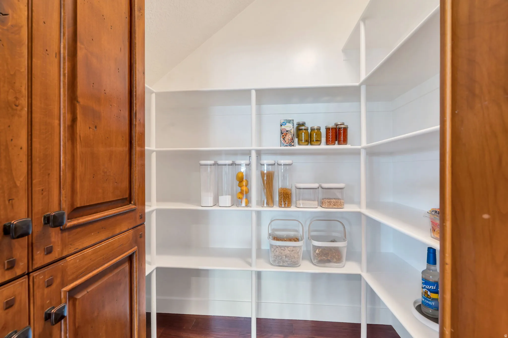 Spacious pantry featuring extensive built-in white shelving, wood-finish flooring, and natural wood cabinetry with dark hardware