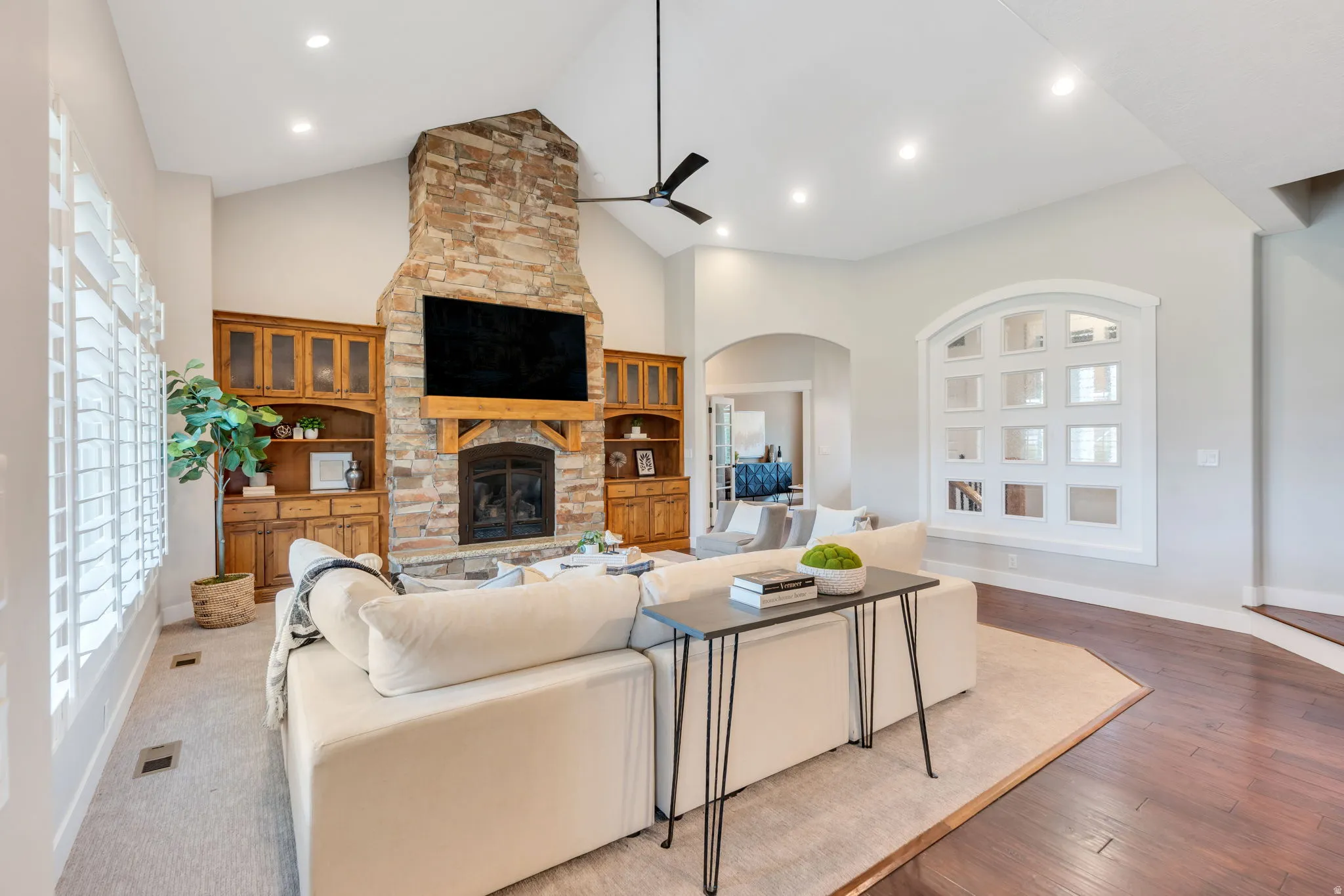 Vaulted ceiling living area featuring a stacked stone fireplace with wood mantel, built-in custom cabinetry, recessed lighting, and plantation shutters