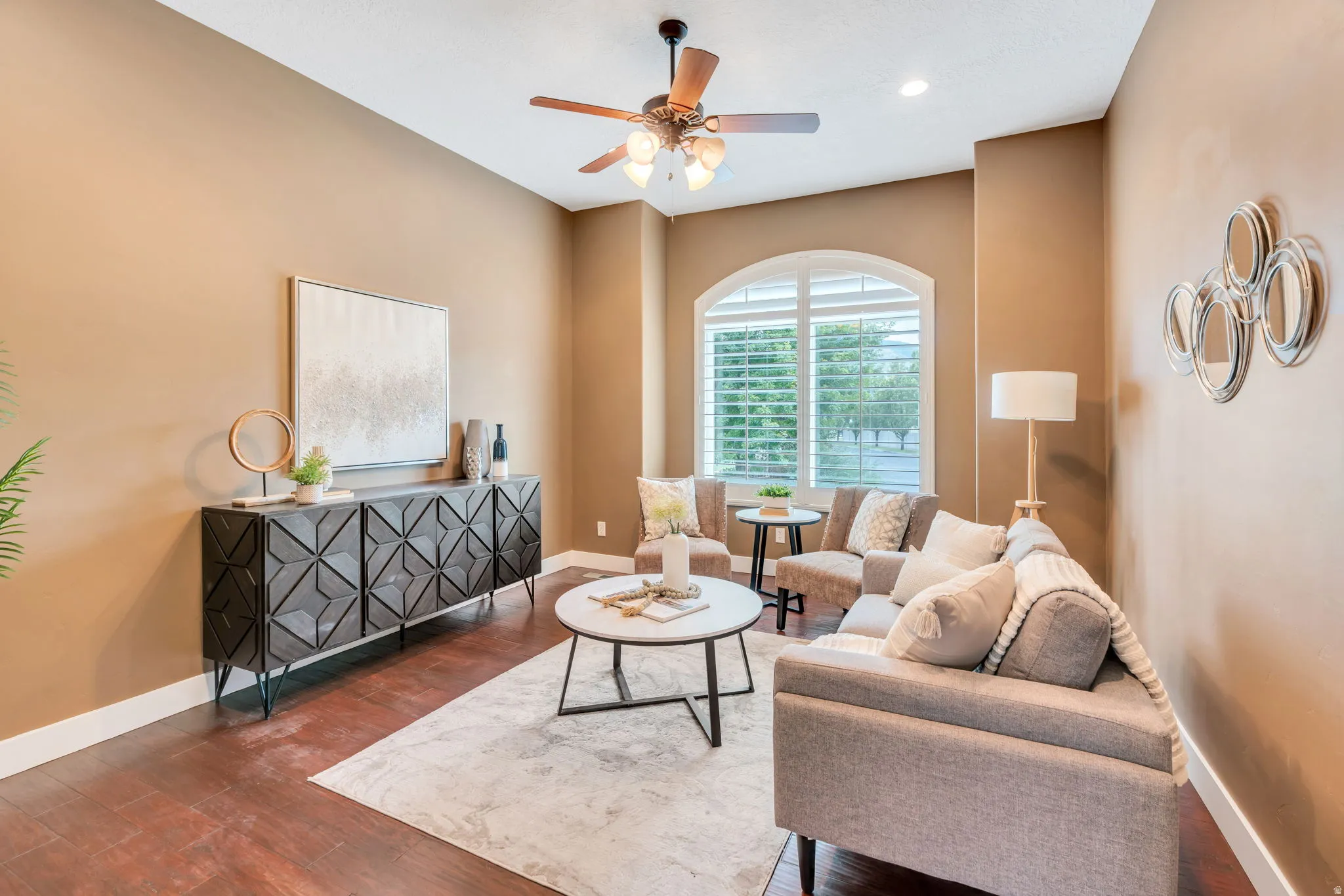Living area featuring wood-finish flooring, an arched window with louvered shutters, a ceiling fan with integrated lighting, and crown molding
