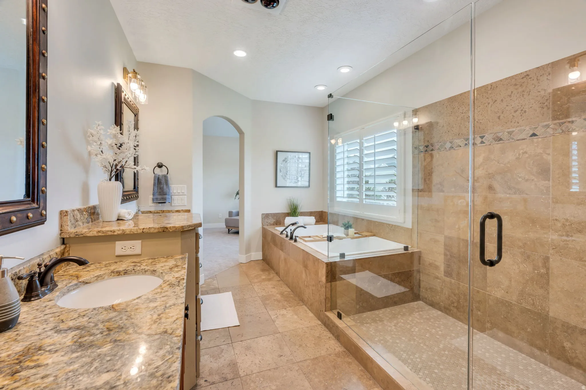Spacious bathroom featuring a built-in tub, separate glass-enclosed shower, dual vanity with stone countertops, recessed lighting, and plantation shutters