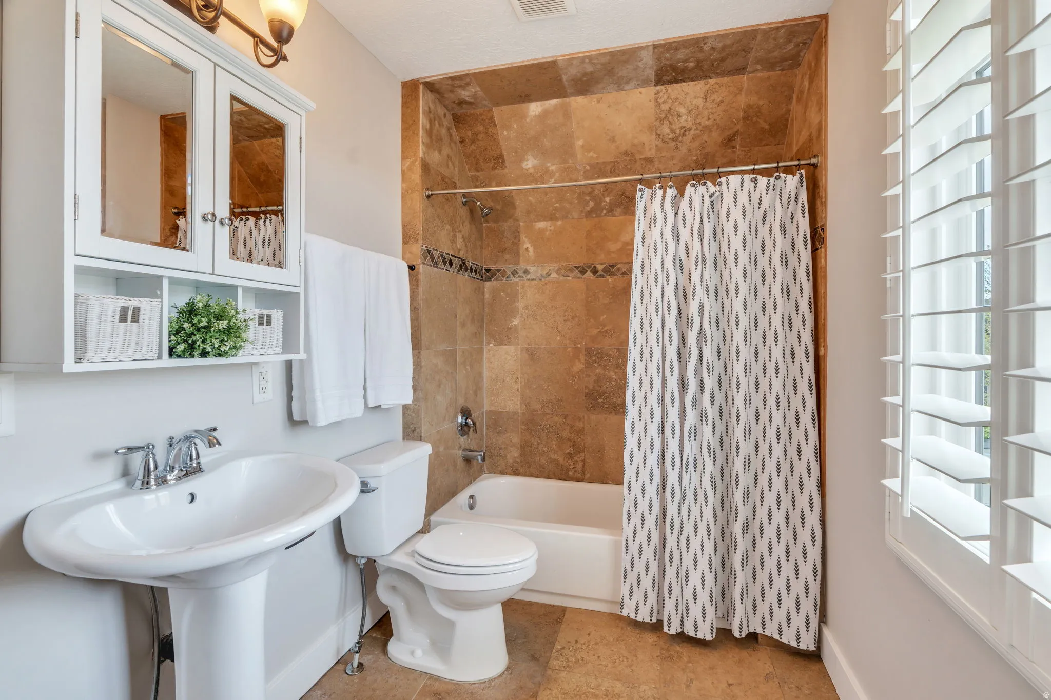 Pedestal sink with chrome faucet, white medicine cabinet with integrated shelving, tiled tub surround with decorative accent, white bathtub, and plantation shutters