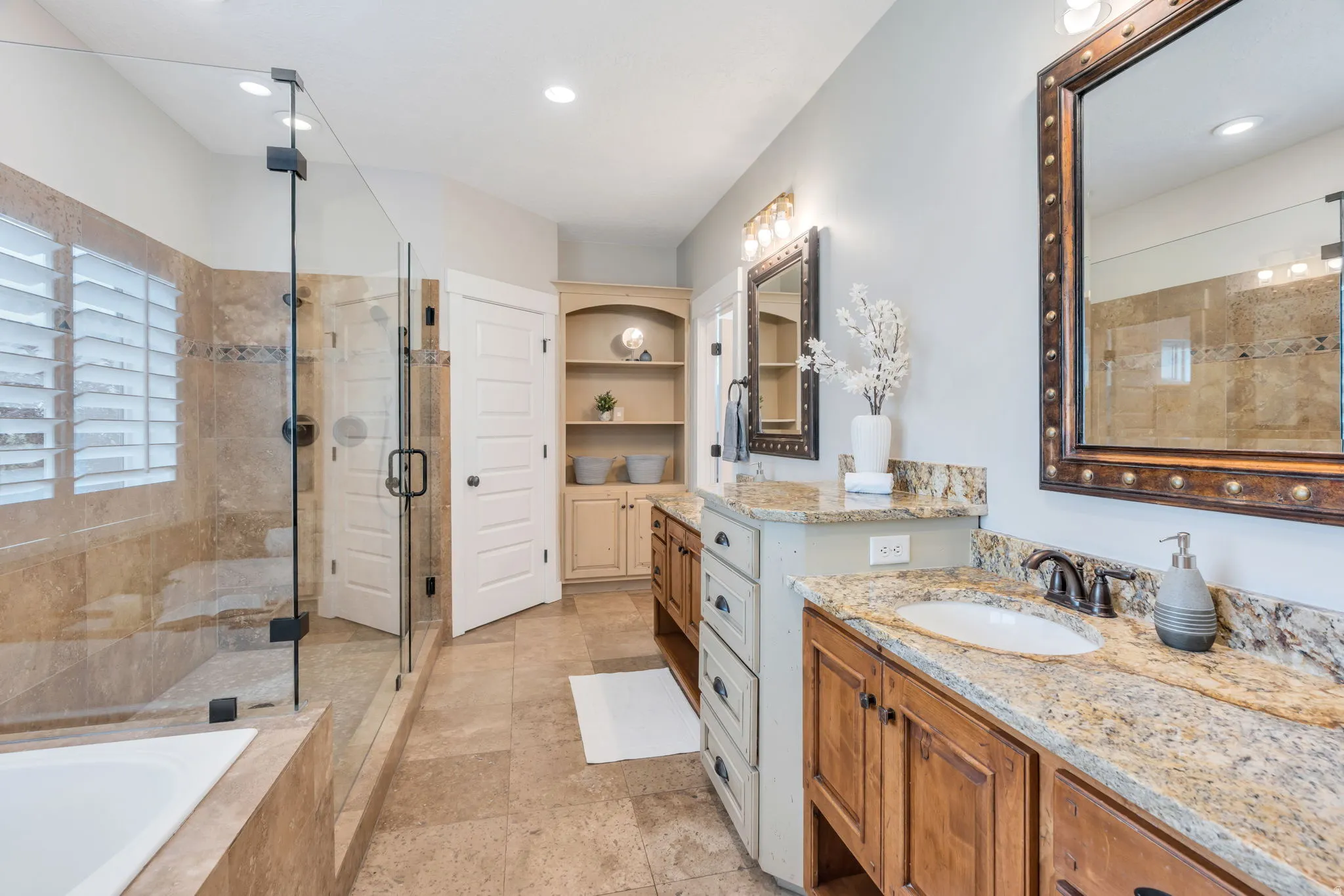 Tiled walk-in shower with a glass enclosure, soaking tub, dual vanity with granite countertops, and built-in shelving