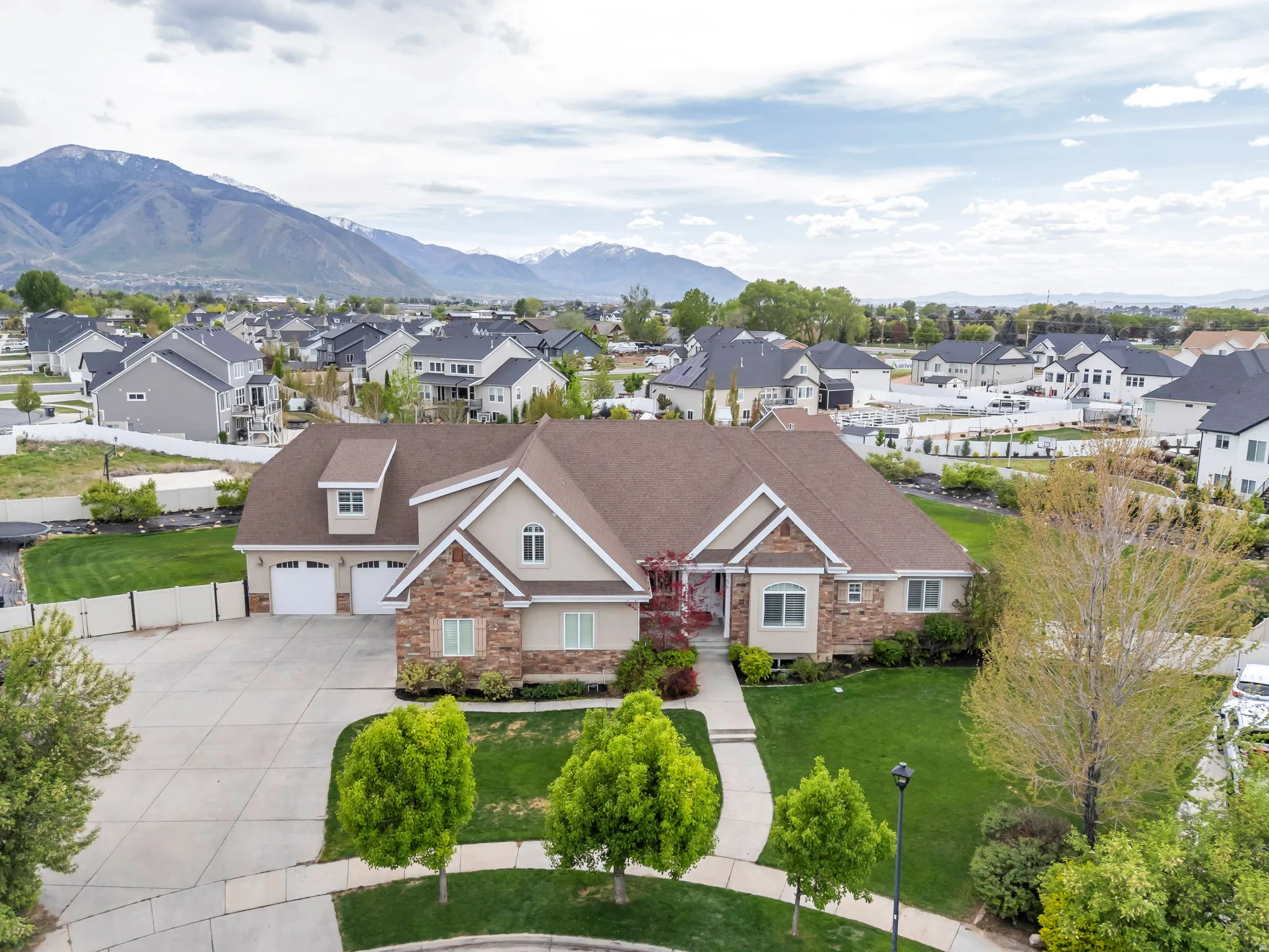 Expansive residence featuring a multi-gable roofline, stone and stucco exterior, three-car garage, established front yard landscaping, and a concrete driveway with an integrated walkway