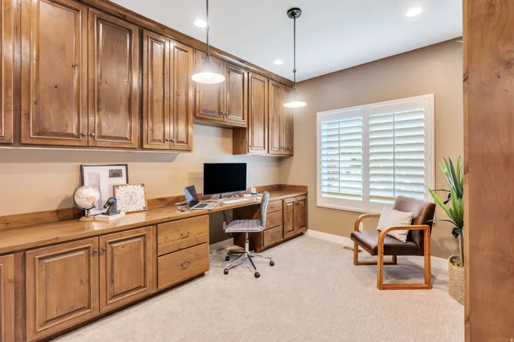 Dedicated office space featuring extensive wood cabinetry and integrated desk, pendant lighting, recessed ceiling lights, and window with plantation shutters