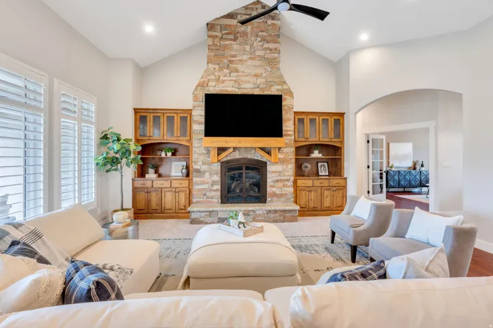 Vaulted ceiling living area featuring a stacked stone fireplace with wood mantel, built-in cabinetry, recessed lighting, and plantation shutters