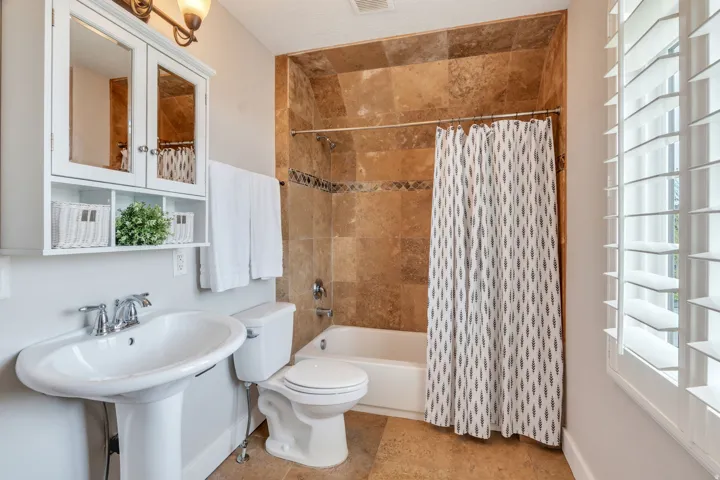 Pedestal sink with chrome faucet, white medicine cabinet with integrated shelving, tiled tub surround with decorative accent, white bathtub, and plantation shutters