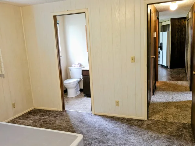 Unfurnished bedroom featuring wooden walls, dark carpet, a textured ceiling, ensuite bathroom, and white refrigerator with ice dispenser