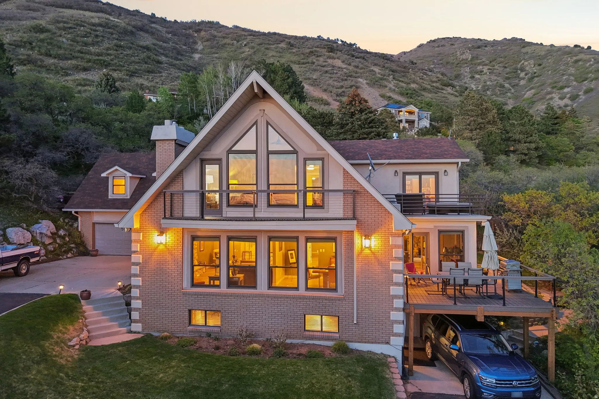 Back of house at dusk featuring a chimney, brick siding, a balcony, a mountain view, and a lawn