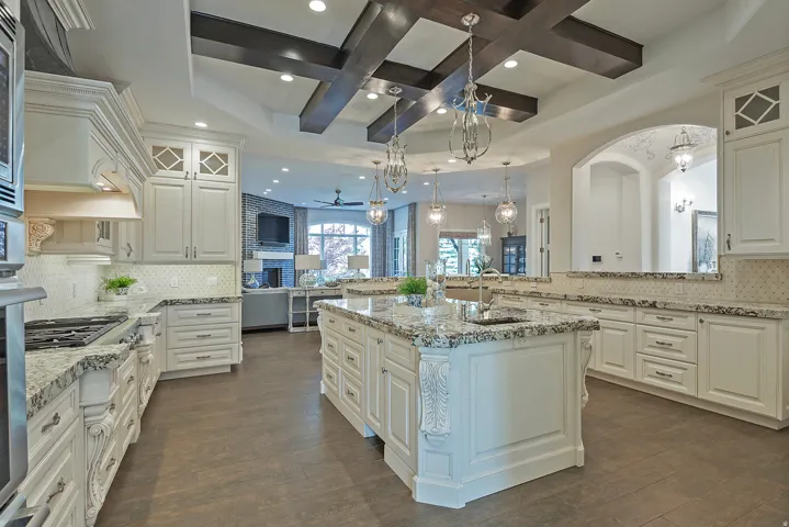 Kitchen featuring backsplash, glass insert cabinets, coffered ceiling, a center island with sink, and light stone counters