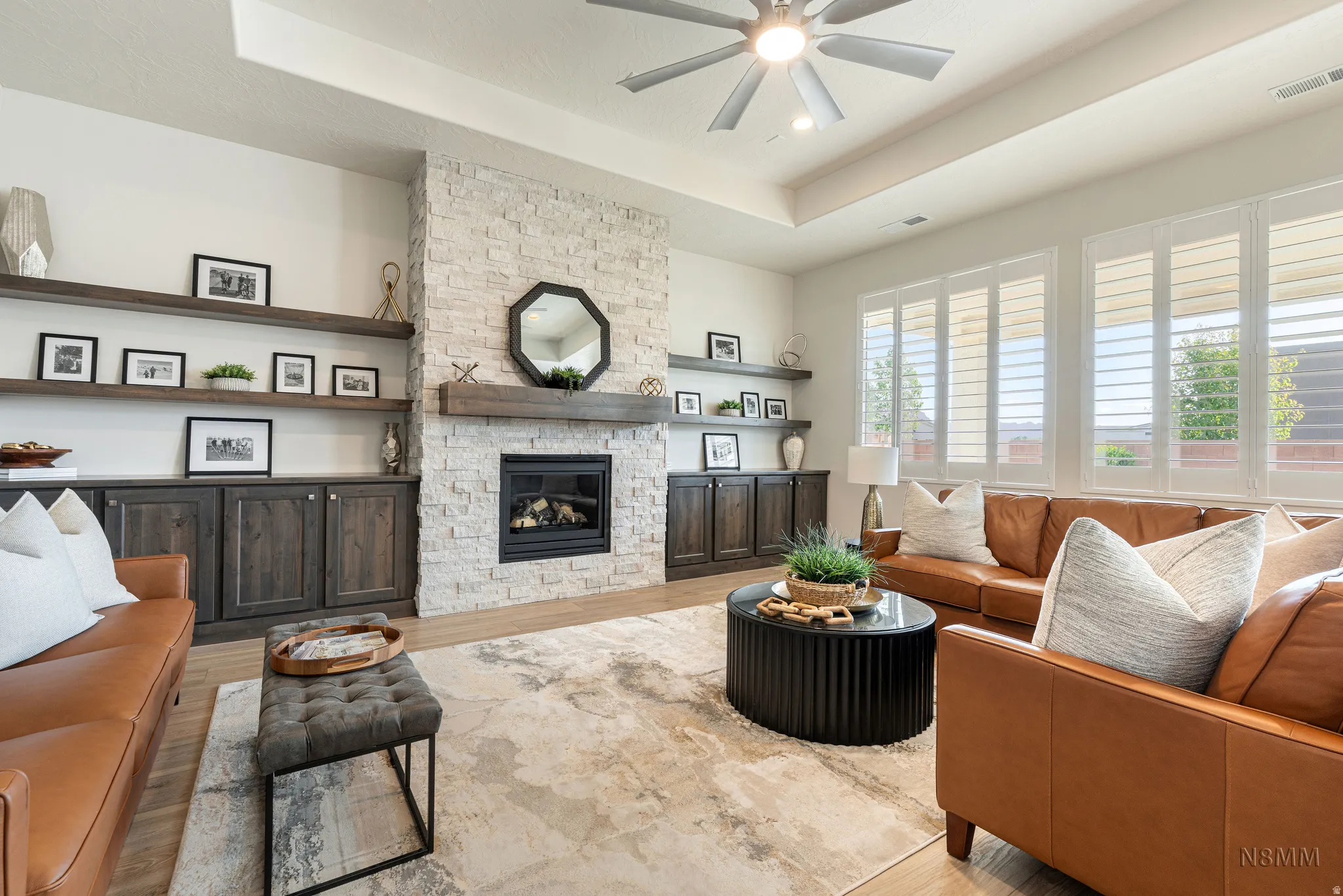 Living room featuring a tray ceiling, wood finished floors, ceiling fan, and a stone fireplace