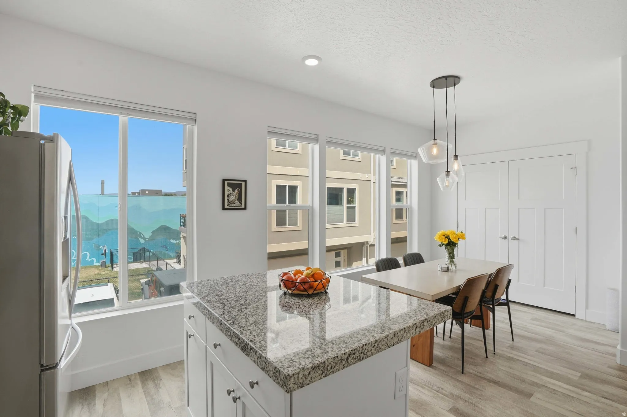 Kitchen featuring stainless steel fridge with ice dispenser, light wood-type flooring, hanging light fixtures, a center island, and light stone counters