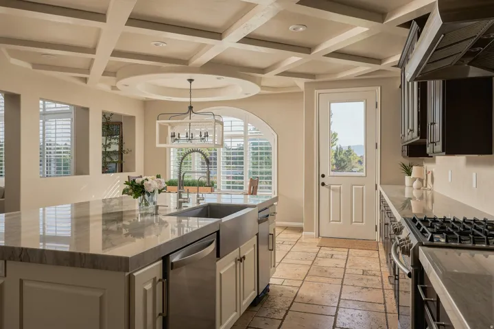 Coffer ceiling with recessed lighting, central island with an undermount sink, stainless steel appliances, stone tile flooring, and custom cabinetry
