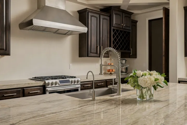 Expansive kitchen island, stainless steel range hood, built-in wine rack, dark wood cabinetry, and a double basin sink with a gooseneck faucet