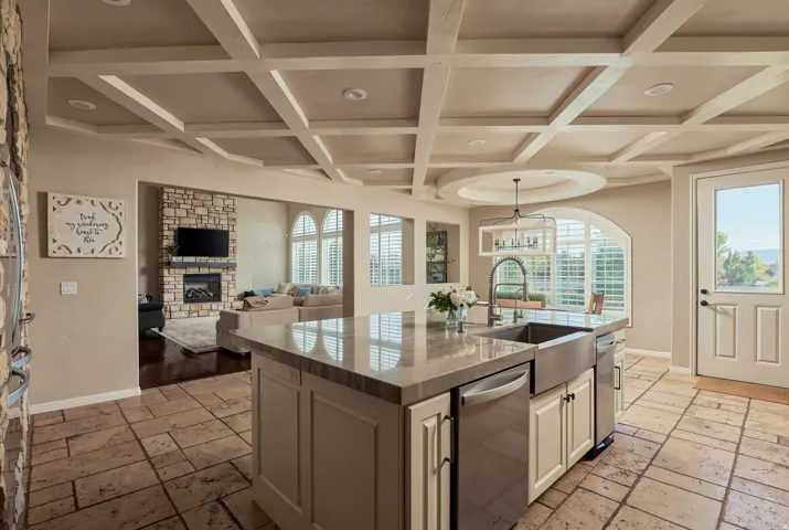 Coffered ceiling with recessed lighting, stone tile flooring, central island with integrated stainless steel appliances, stone-clad fireplace, and arched windows with plantation shutters