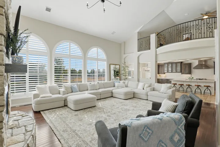 Expansive living area with high ceilings, arched windows with plantation shutters, a stone-clad fireplace with a wood mantel, and wood-finish flooring