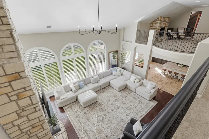 Expansive living space with vaulted ceilings, arched windows featuring plantation shutters, a stone-clad fireplace, and a second-story balcony with decorative wrought iron railing