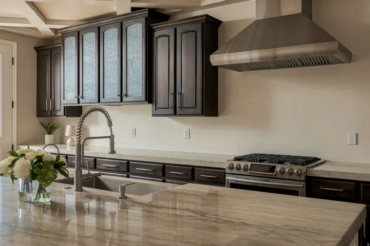 Dark wood cabinetry with textured glass inserts, stainless steel range hood, stainless steel gas range, stone countertops, and a gooseneck faucet
