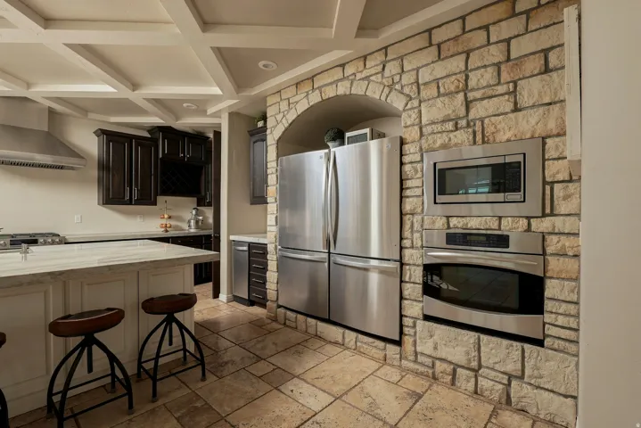 Coffered ceiling, stone-clad archway, integrated stainless steel appliances, dark wood-finish cabinetry, and stone tile flooring